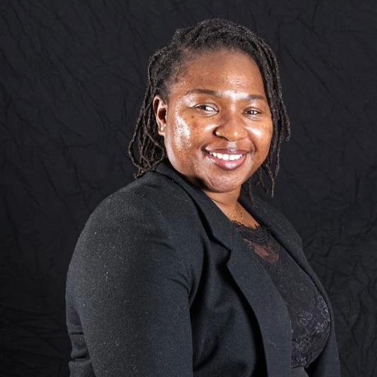 Professional portrait of a smiling woman with dreadlocks, wearing a black blazer against a dark background.