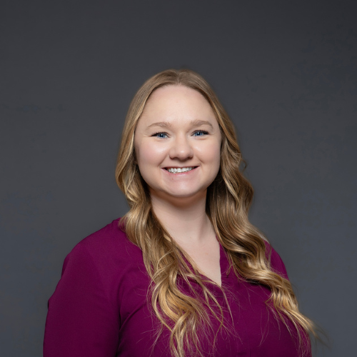 Professional headshot of a woman with long, wavy hair wearing a burgundy top, set against a gray background.